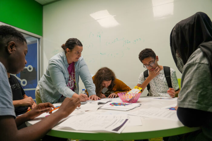 A UVA teacher assists four UVA students at a round table in a classroom with math equations on the whiteboard behind them.
