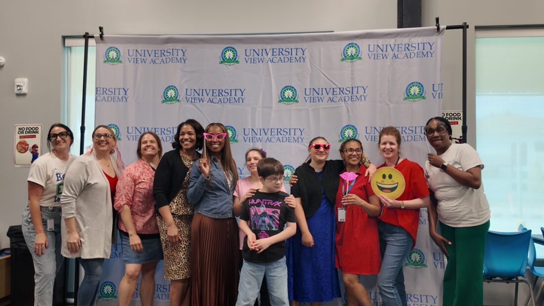 Smiling teachers and students in front of a University View Academy banner, some wearing sunglasses and holding playful props.