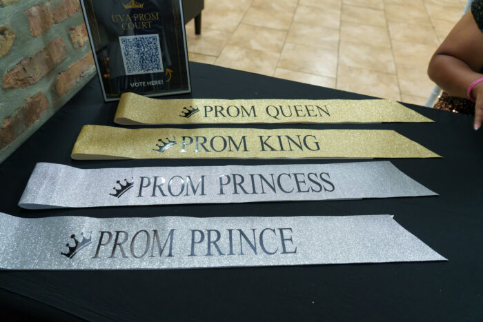 Four glittery sashes reading Prom Queen, Prom King, Prom Princess, and Prom Prince are laid out on a black table.