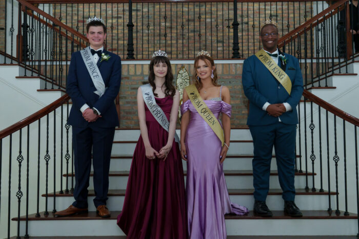 Four people stand on stairs, each wearing crowns and sashes labeled prom king, queen, prince, and princess.