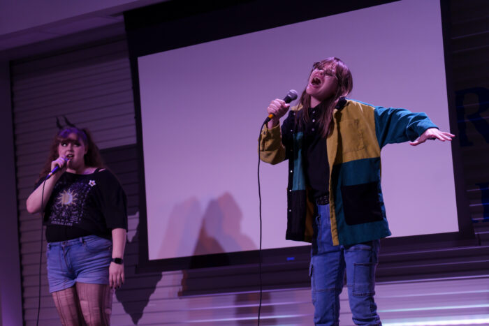 A student with her arms outstretched sings passionately into a microphone on stage.