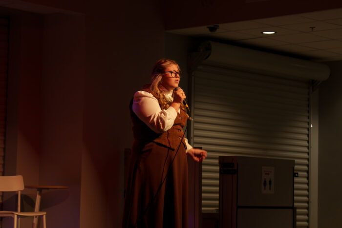 A student in glasses and a brown dress sings into a microphone on stage.