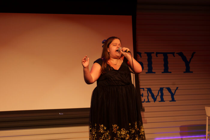 A student in a black dress sings into a microphone on stage.