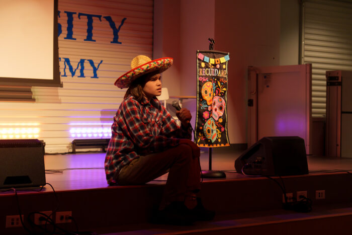 A student wearing a sombrero sits on stage and sings into a microphone beside a vibrant Día de los Muertos banner.