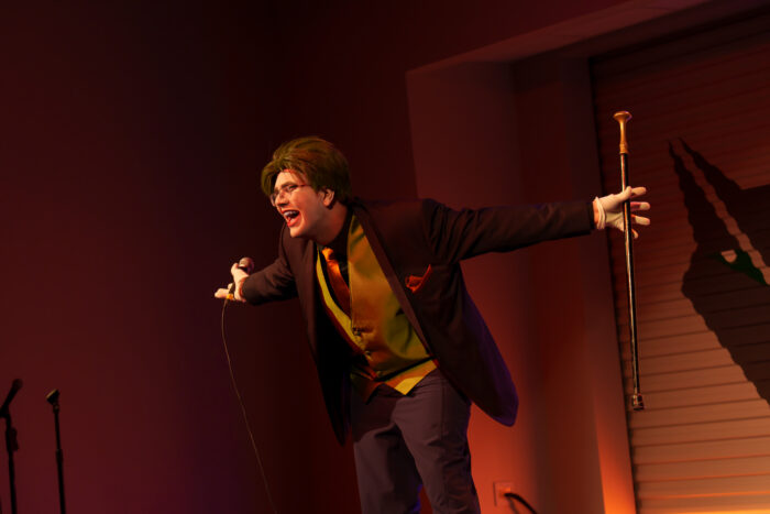 A student dressed in a suit with a green wig, holding a cane and microphone, leans forward on stage under dramatic lighting.