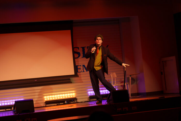 A student dressed as the Joker holds a microphone and twirls a cane, posing on stage.