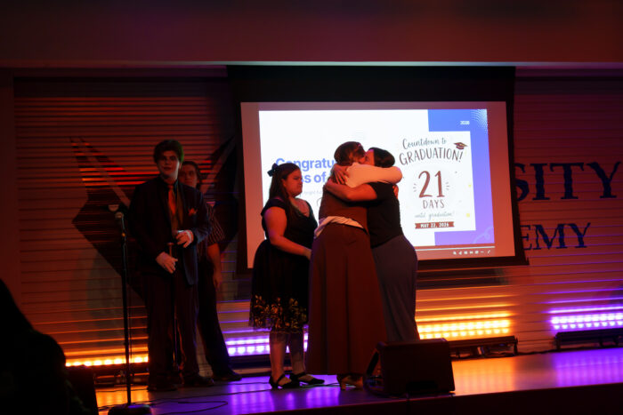 Several students stand on stage with a graduation slideshow in the background. One student embraces her teacher.