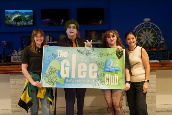 Glee club students, one in costume, hold up a banner for the glee club and smile for the camera.