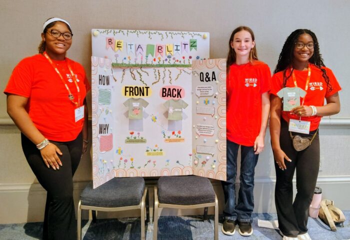 Three students stand beside a project board display with diagrams.