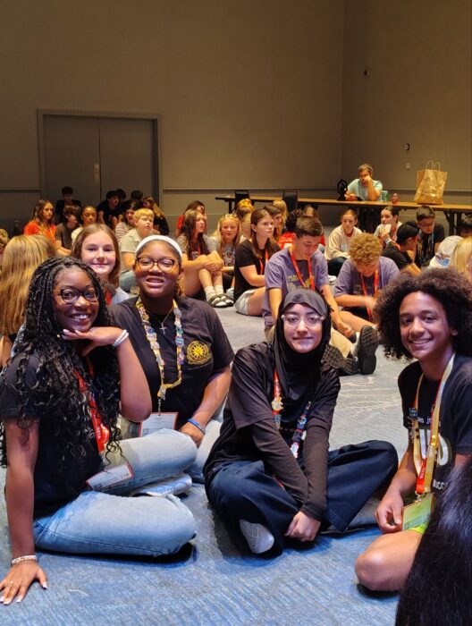 Students sit cross-legged on a carpeted floor at an indoor event, with others seated in the background.