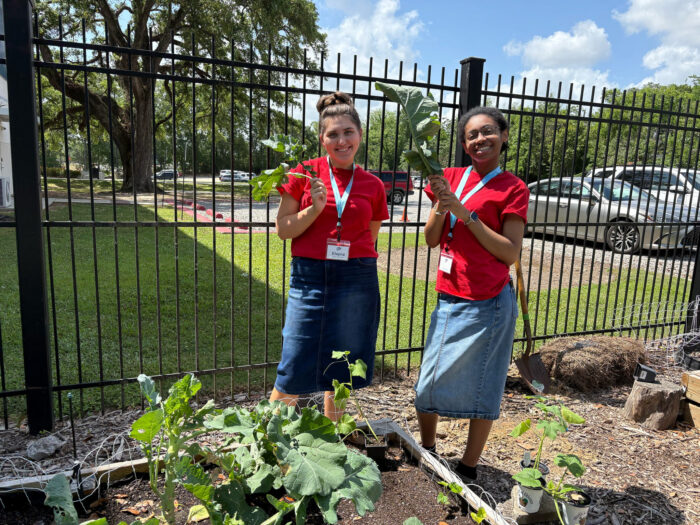Two students stand in a garden, smiling and holding large green leaves near a fence.