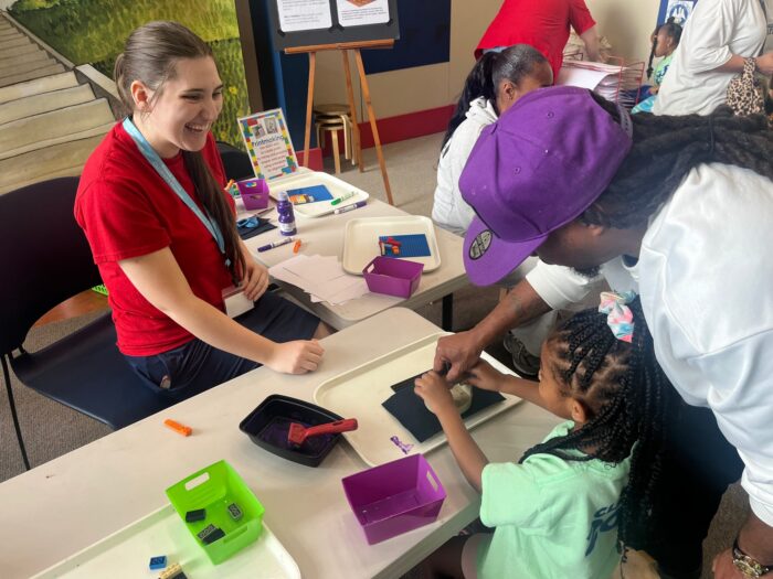 A young girl works on a craft at a table with an adult as a smiling student intern watches. Craft supplies fill the table.