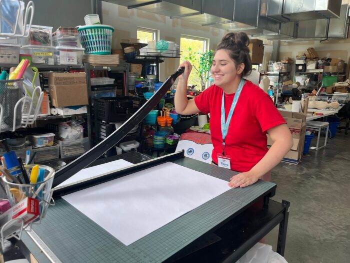 A smiling student uses a large paper cutter in an art supply room with shelves and boxes.