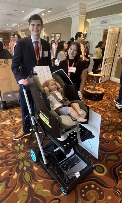 Two students stand beside a baby doll in a high-tech car seat at an indoor conference.