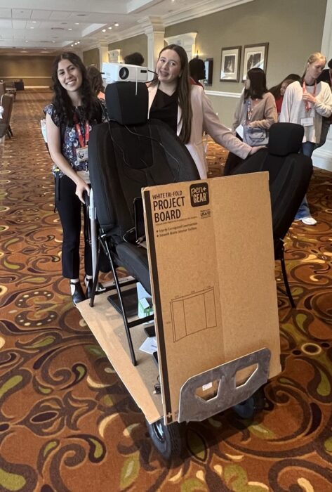 Two students stand behind a cart with a car seat and a project board in a carpeted conference room with others nearby.