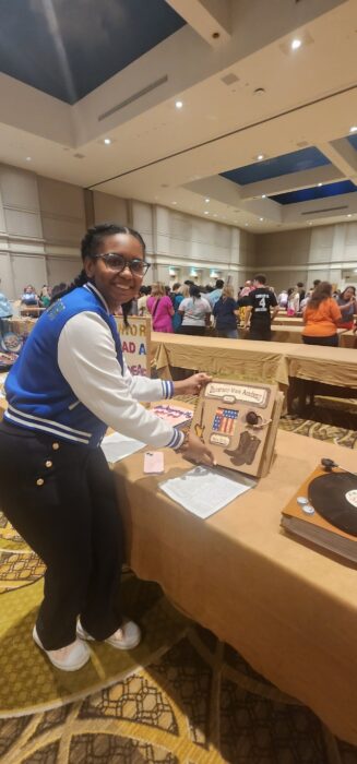 A student stands by a decorated project display at an indoor event with other attendees.