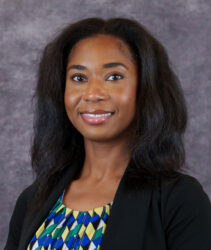 Brielle Jones headshot. Smiling woman with long dark hair, patterned top, and black blazer stands before a gray mottled background.