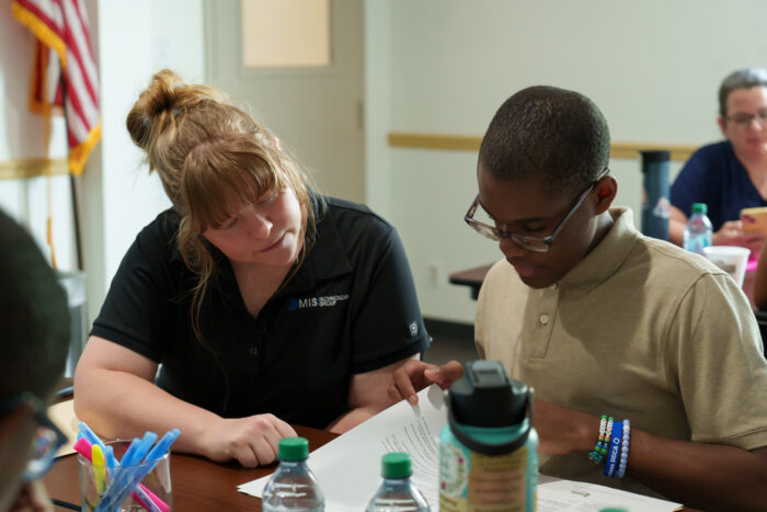 A woman and boy sit at a table looking at papers, with water bottles, pens, and other people in the background.