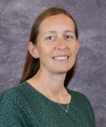 Elizabeth Alexander. A smiling woman with long brown hair and a green patterned top stands before a gray textured background.