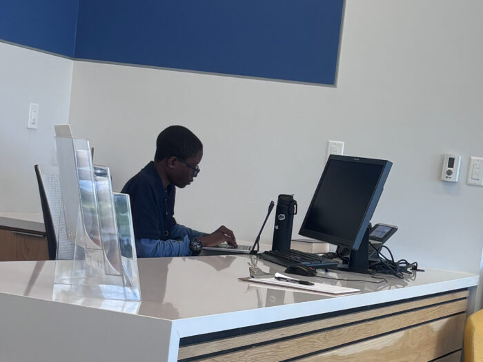 Student at a desk using a laptop, with a desktop monitor, water bottle, and office supplies close by.