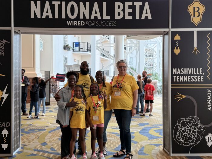 Six people pose beneath a “National Beta” sign at an indoor event.