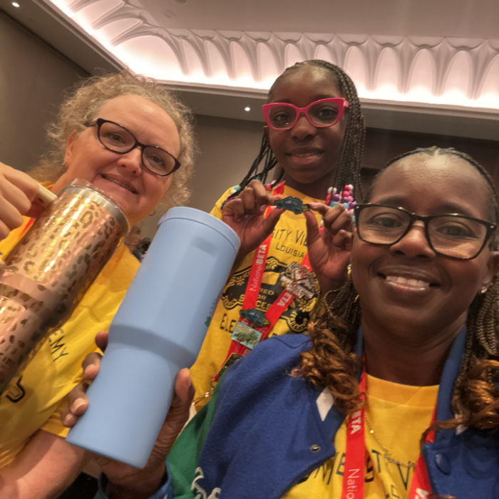 A student and two teachers in yellow shirts and lanyards pose for a selfie at an indoor event, holding tumblers and a pin.