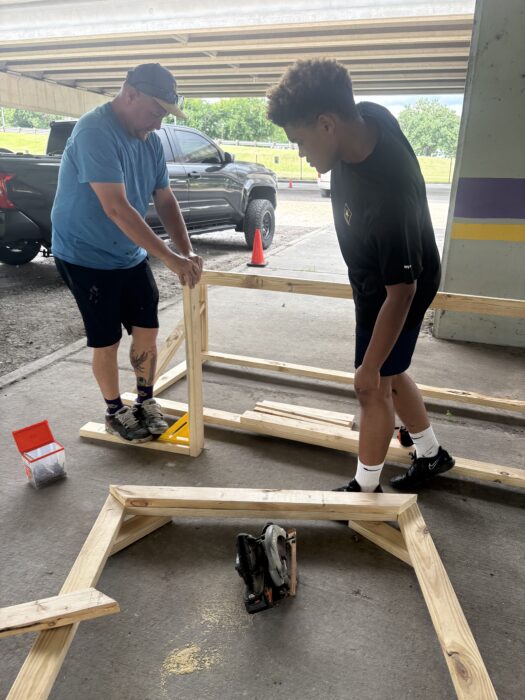Two people build a wooden frame on the ground near tools under a parking garage, with a truck and orange cone close by.