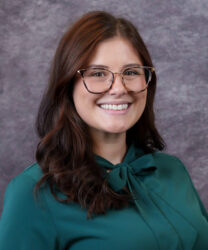 Jenni Falcon headshot. Smiling woman with long brown hair, glasses, and a green blouse in front of a gray textured background.