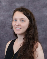 Megan O'Dell headshot. Woman with long wavy brown hair in a black sleeveless top stands before a textured gray background.