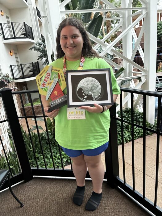 A student stands on a balcony holding a trophy and framed picture.