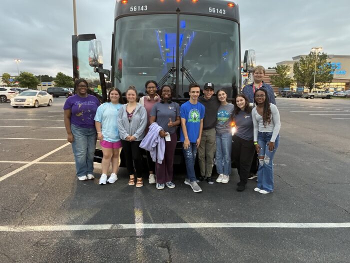 A group of students pose together in front of a parked bus in a parking lot.