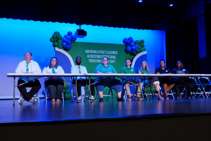 A panel of teachers sit at a table on stage.