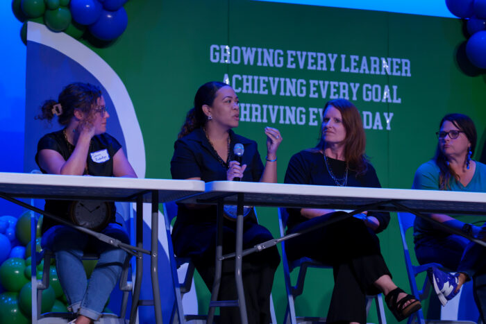 A teacher speaks into a microphone at a panel table seated with other panelists.