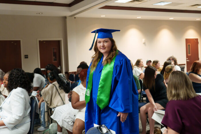 A graduate wearing a blue cap and gown with a green stole walks down an aisle among seated guests at an indoor ceremony.