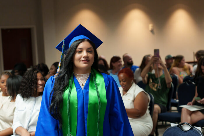 A graduate in a blue cap and gown with a green stole stands walks down an indoor aisle as an audience watches in the background.