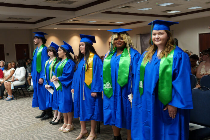 Six graduates in blue caps and gowns with green sashes stand in a row indoors, while an audience watches.