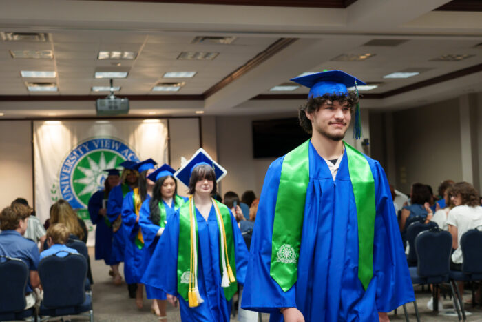 Graduates in blue gowns and green stoles walk down the aisle at a decorated hall during a graduation ceremony.