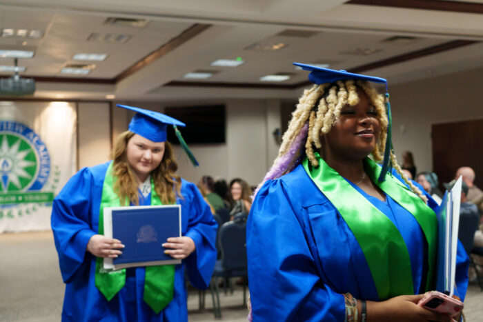 Two graduates in blue gowns and green stoles hold diplomas and exit the ceremony.