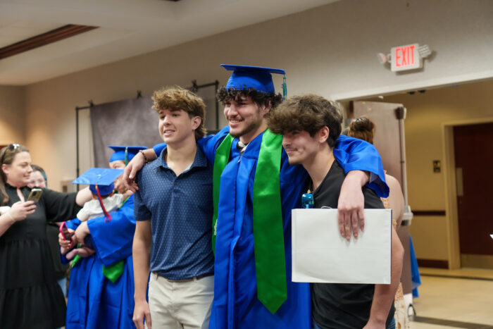 Three students pose at a graduation ceremony, one in a blue cap and gown smiles with his arms around the others.