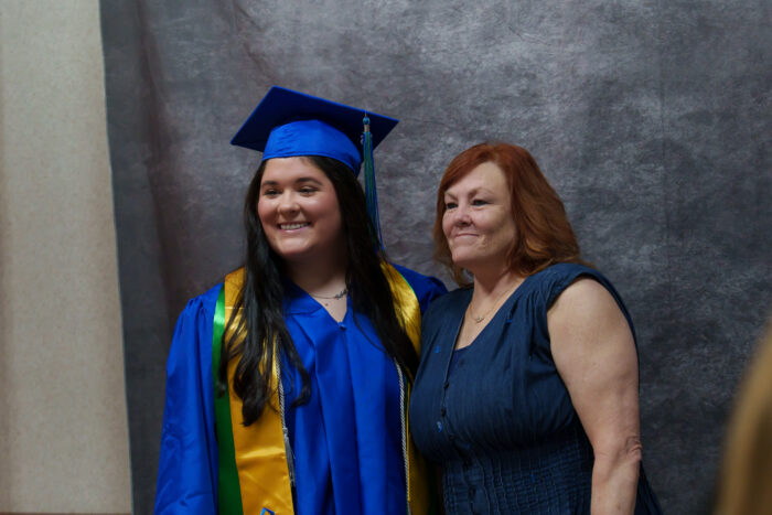 A graduate and another woman pose for a picture against a gray backdrop.
