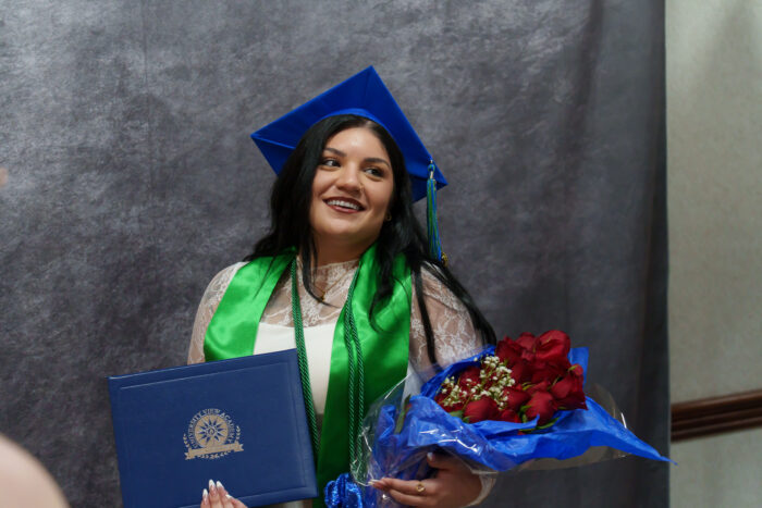 Smiling student in a graduation cap and gown holds a diploma and red roses against a gray background.