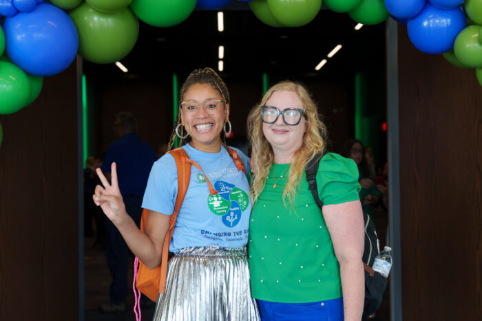 Two staff members smile for a picture under green and blue balloons.