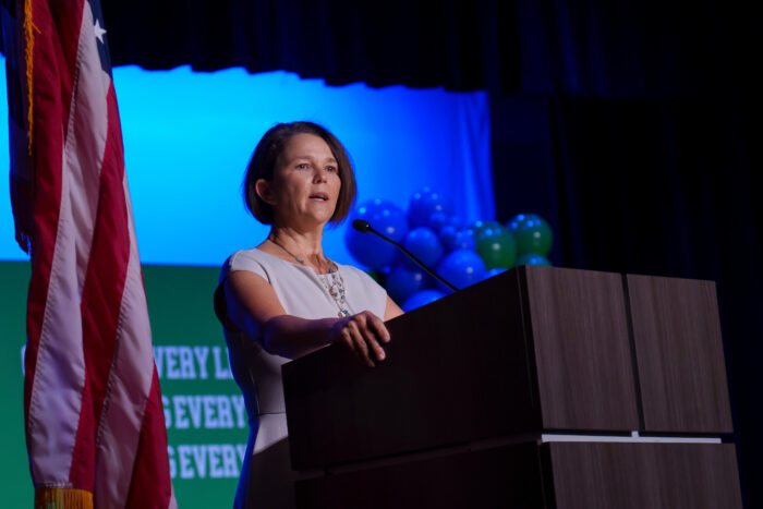Caroline Roemer speaks at a podium on stage, with an American flag and balloons visible in the background.