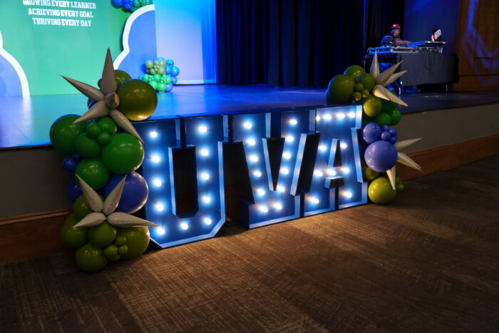 Illuminated “UVA” sign decorated with green, blue, and white balloons in front of a stage.