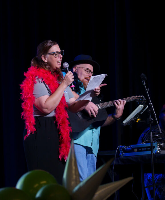 A teacher in a red feather boa sings into a microphone while another plays guitar.