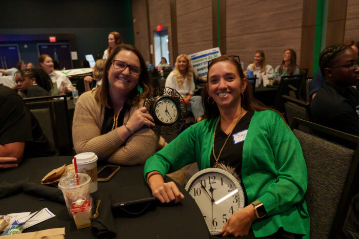 Two seated teachers holding clocks smile for a picture.