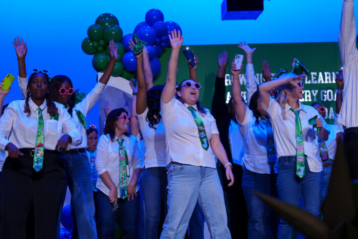 Several teachers in white shirts, green ties, and sunglasses dance on stage.