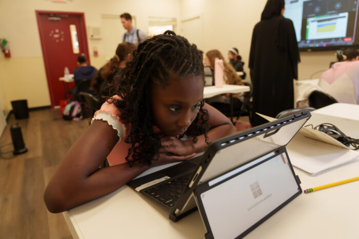 A student studies a laptop at her desk, while classmates and a teacher are in the background.