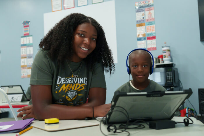 An adult and a child sit at a table with a laptop. The child wears headphones, and they both smile at the camera.