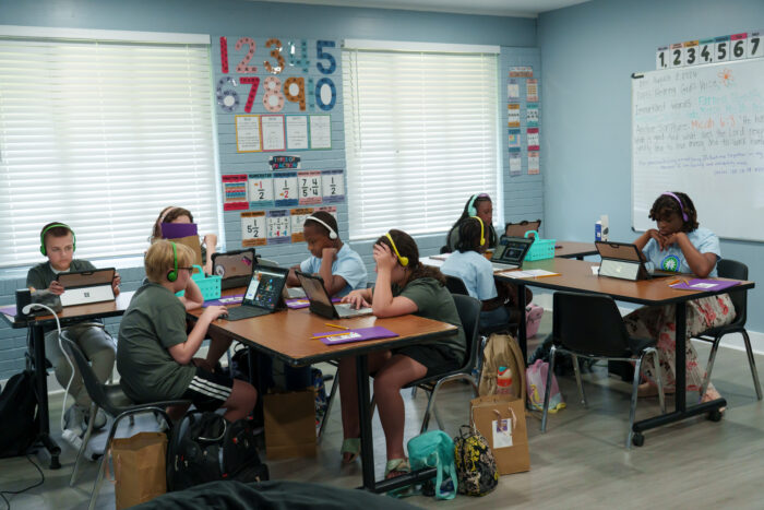 Students wearing headphones use laptops at tables in a classroom.